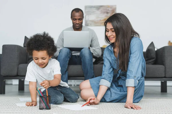 Afro-américaine mère et fils dessin sur le sol à la maison, père travaillant avec ordinateur portable — Photo de stock