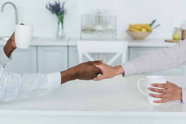 Cropped image of african american girlfriend and boyfriend holding hands and cups of coffee at home — Stock Photo