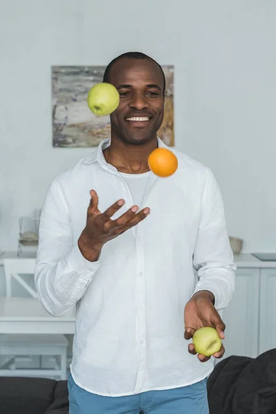 Guapo afroamericano hombre malabares con frutas en casa - foto de stock