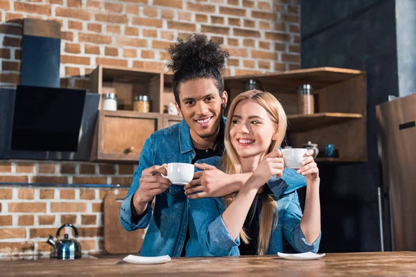 Retrato de casal jovem multirracial sorridente com xícaras de café na cozinha em casa — Fotografia de Stock