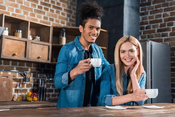 Retrato de casal jovem multirracial sorridente com xícaras de café na cozinha em casa — Fotografia de Stock