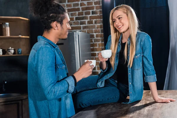 Sonriente pareja joven multirracial con tazas de café teniendo conversación en la cocina en casa - foto de stock