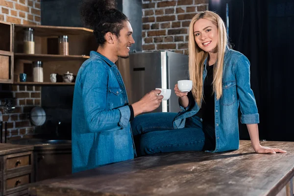 Sonriente pareja joven multirracial con tazas de café en la cocina en casa - foto de stock