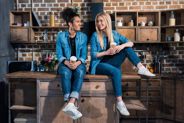 Улыбающаяся многорасовая молодая пара с кофе, чтобы смотреть друг на друга на кухне дома — стоковое фото