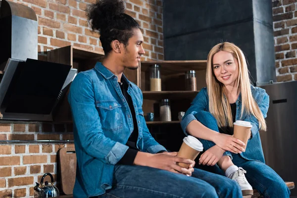 Multirassische junge Paar mit Kaffee zu gehen auf dem Tresen in der Küche zu Hause sitzen — Stockfoto