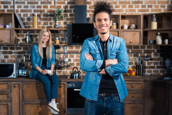 Foco seletivo de homem americano africano sorridente com namorada caucasiana sentada no balcão atrás na cozinha — Fotografia de Stock