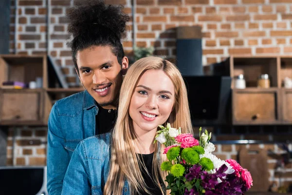 Retrato de mulher caucasiana feliz com buquê de flores e namorado afro-americano por perto na cozinha em casa — Fotografia de Stock