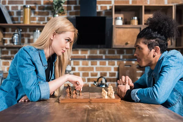 Вид сбоку на многонациональную молодую пару, играющую вместе в шахматы на кухне — стоковое фото