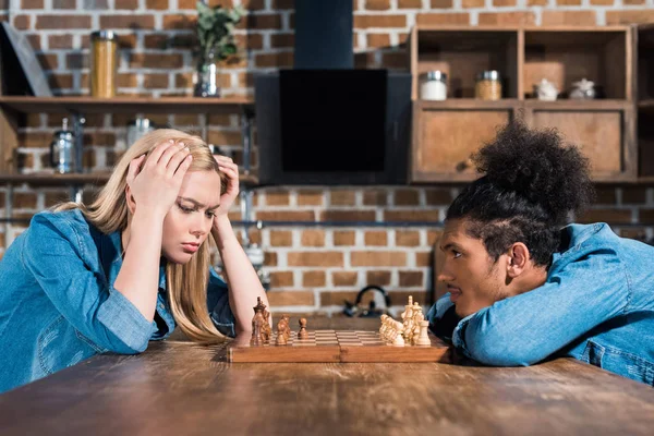 Вид сбоку на многонациональную молодую пару, играющую вместе в шахматы на кухне — стоковое фото