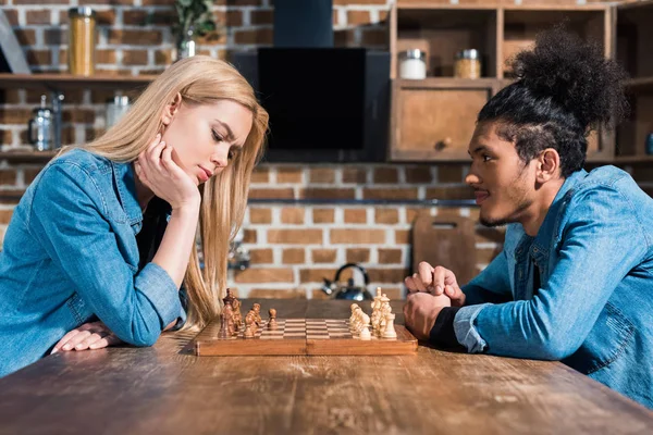 Vue de côté de jeune couple multiethnique jouant aux échecs ensemble dans la cuisine — Photo de stock