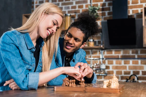 Улыбающаяся многонациональная молодая пара, играющая в шахматы на кухне — стоковое фото