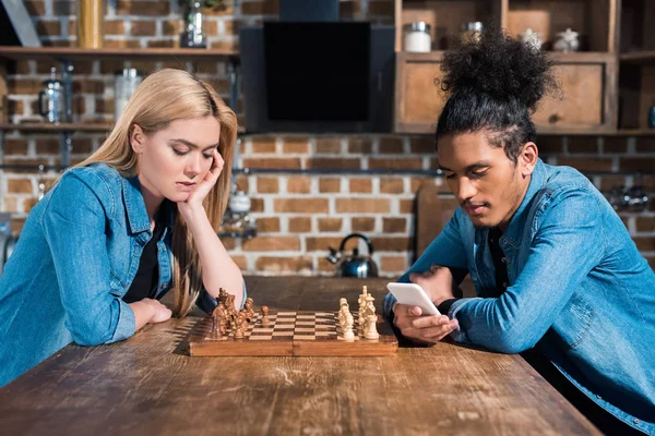 Vista lateral de mujer caucásica aburrida y hombre afroamericano con teléfono inteligente en la mesa con ajedrez en la cocina - foto de stock