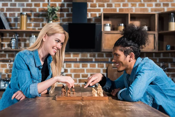 Vista lateral de sonriente pareja joven multiétnica jugando ajedrez juntos en la cocina - foto de stock