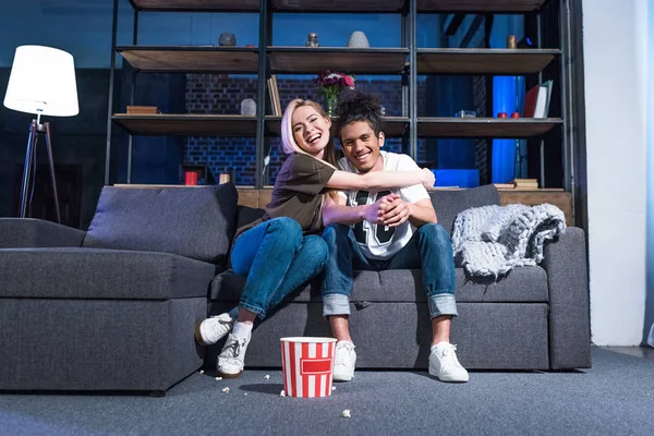 Sonriente pareja multirracial con palomitas de maíz viendo película en el sofá juntos en casa - foto de stock