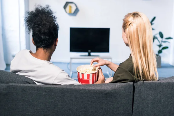 Vista trasera de la pareja multirracial con palomitas de maíz viendo películas juntos en casa - foto de stock