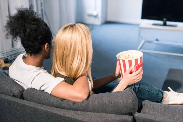 Visão traseira do casal multirracial com pipocas assistindo filme juntos em casa — Fotografia de Stock