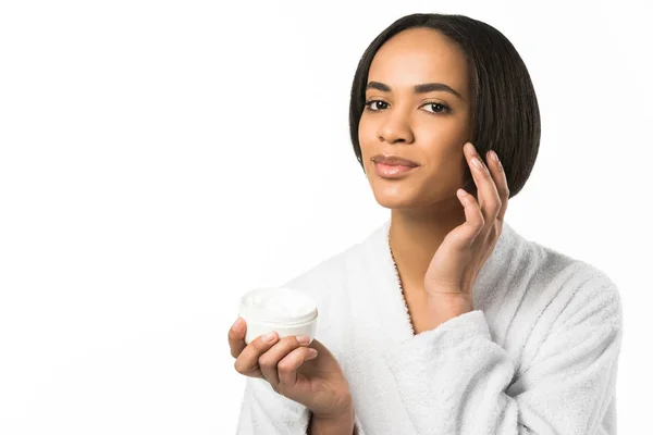 Donna afroamericana in accappatoio che applica crema per la pelle sul viso, isolata su bianco — Foto stock