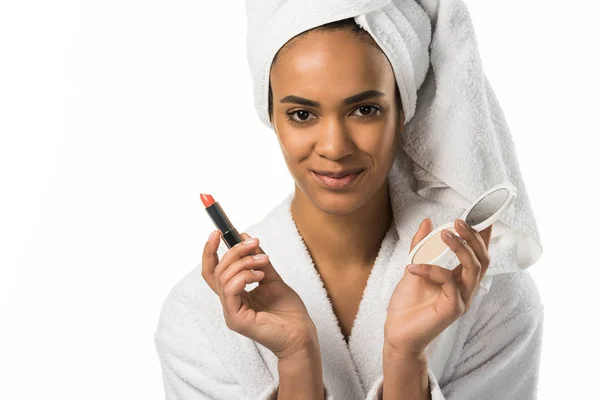 Belle femme afro-américaine en peignoir et serviette tenant rouge à lèvres et miroir, isolée sur blanc — Photo de stock