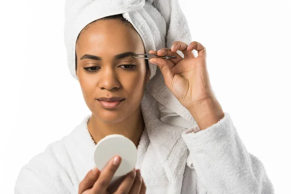 Mujer afroamericana mirando al espejo y arrancando cejas con pinzas, aislada en blanco - foto de stock