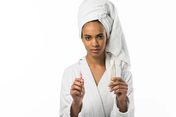 Chica afroamericana en albornoz y toalla sosteniendo pasta de dientes y cepillo de dientes, aislada en blanco - foto de stock
