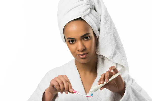 Femme afro-américaine en peignoir et serviette avec dentifrice et brosse, isolée sur blanc — Photo de stock