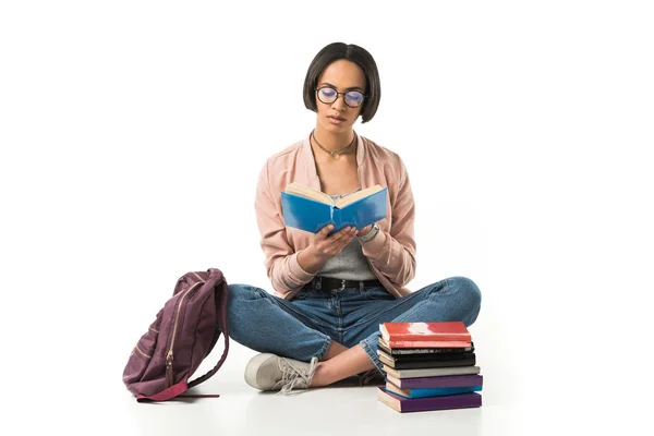 Magnifique étudiant afro-américain lisant des livres assis sur le sol avec sac à dos, isolé sur blanc — Photo de stock