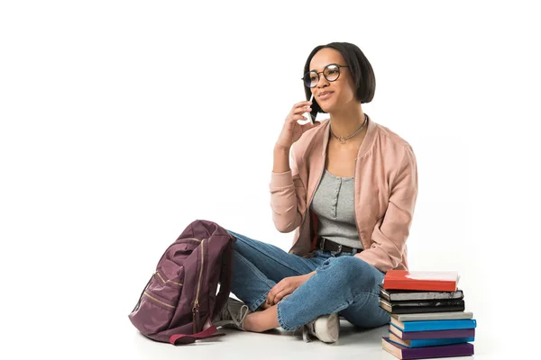 Estudiante afroamericana hablando por teléfono inteligente y sentada con mochila y libros en el suelo, aislada en blanco - foto de stock