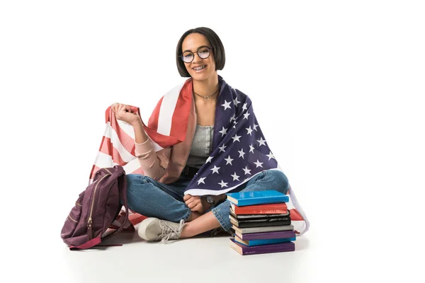 Estudante afro-americano feminino envolto em bandeira dos eua sentado no chão com mochila e livros, isolado em branco — Fotografia de Stock