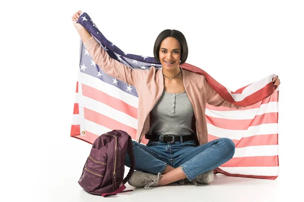 Sonriente estudiante afroamericano sosteniendo bandera de EE.UU. sentado en el suelo con mochila, aislado en blanco - foto de stock