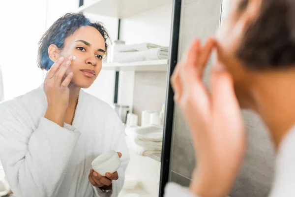 Foco seletivo da menina americana africana aplicando creme hidratante no rosto no banheiro perto do espelho — Fotografia de Stock