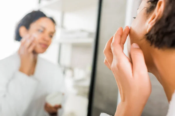 Focalizzazione selettiva della donna afro-americana che applica crema cosmetica sul viso in bagno — Foto stock