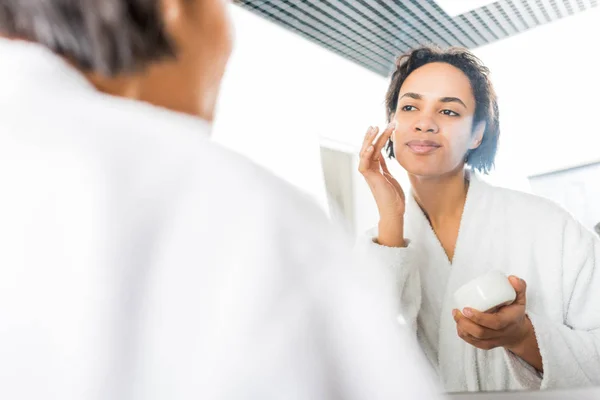 Foco seletivo de mulher afro-americana sorridente aplicando creme facial perto do espelho no banheiro — Fotografia de Stock