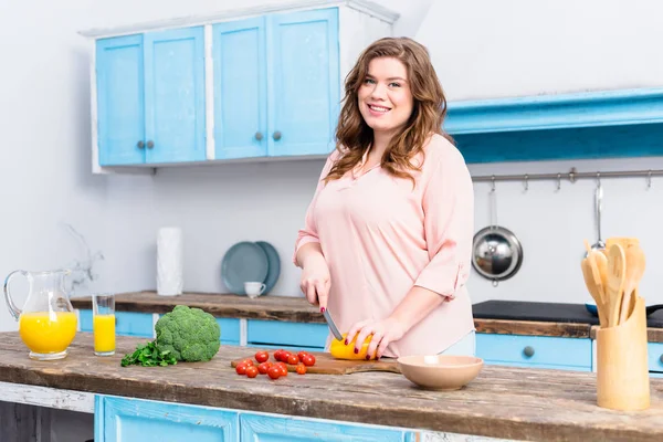 Mujer sonriente joven con sobrepeso cortando verduras para ensalada en la cocina en casa - foto de stock