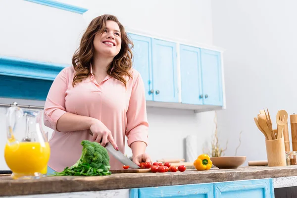 Mujer sonriente joven con sobrepeso cortando verduras para ensalada en la cocina en casa - foto de stock