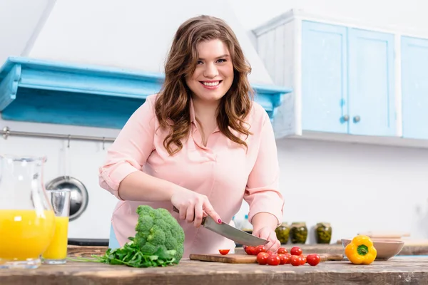 Sovrappeso giovane donna sorridente che taglia verdure per insalata in cucina a casa — Foto stock