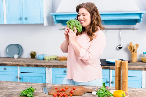 Retrato de mulher sorridente com excesso de peso com brócolis fresco nas mãos na cozinha em casa — Fotografia de Stock