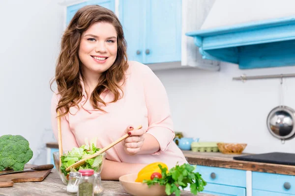 Портрет толстой улыбающейся женщины, смотрящей в камеру во время приготовления свежего салата на ужин на кухне дома — стоковое фото