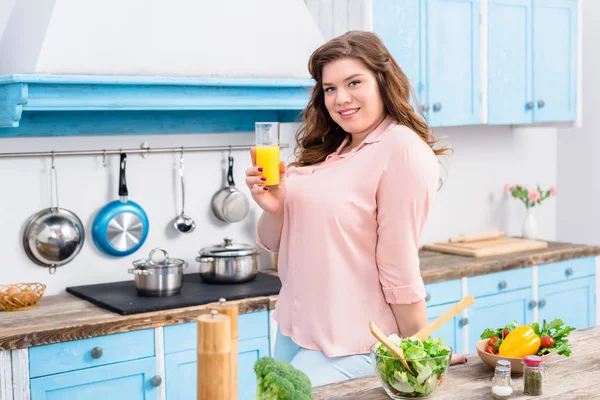 Portrait de femme souriante en surpoids avec verre de jus dans la cuisine à la maison — Photo de stock