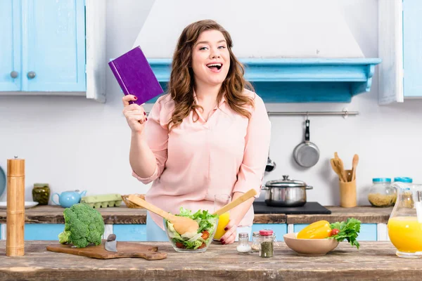 Mujer joven con sobrepeso con libro de cocina de pie en la cocina en casa - foto de stock