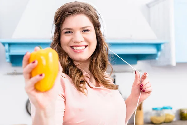 Portrait d'une femme souriante en surpoids dans un casque montrant du poivron frais à la main dans la cuisine — Photo de stock