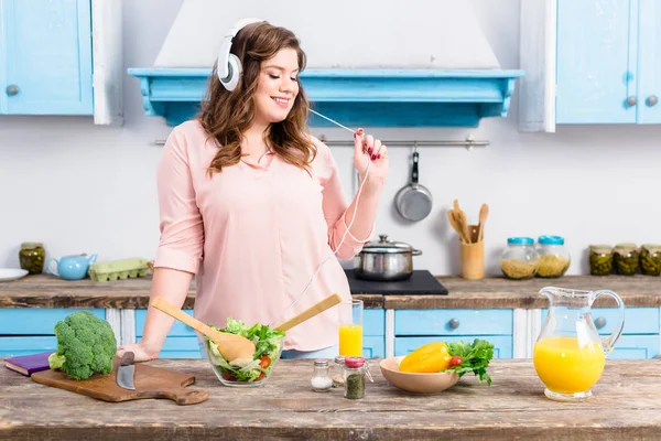 Alegre mujer con sobrepeso escuchando música en auriculares en la mesa con verduras frescas en la cocina en casa — Stock Photo