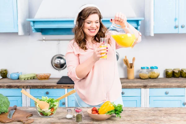 Souriant femme en surpoids dans les écouteurs verser du jus dans le verre dans la cuisine — Photo de stock