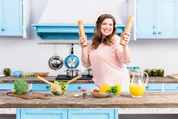 Cheerful overweight woman in headphones with wooden salt and pepper grinders in hands in kitchen — Stock Photo