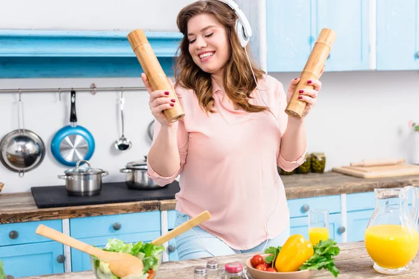 Cheerful overweight woman in headphones with wooden salt and pepper grinders in hands in kitchen — Stock Photo