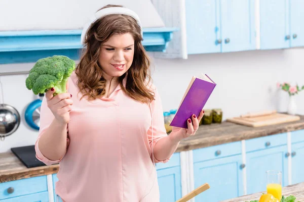 Jeune femme en surpoids dans un casque avec brocoli frais et livre de cuisine dans la cuisine — Photo de stock