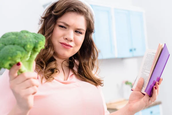 Overweight woman in headphones with cookery book looking at fresh broccoli in hand in kitchen — Stock Photo