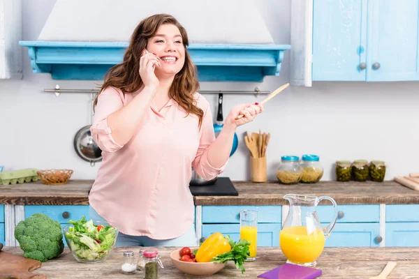 Mujer joven con sobrepeso con cuchara de madera en la mano hablando en el teléfono inteligente mientras está de pie en la mesa con verduras frescas en la cocina en casa - foto de stock