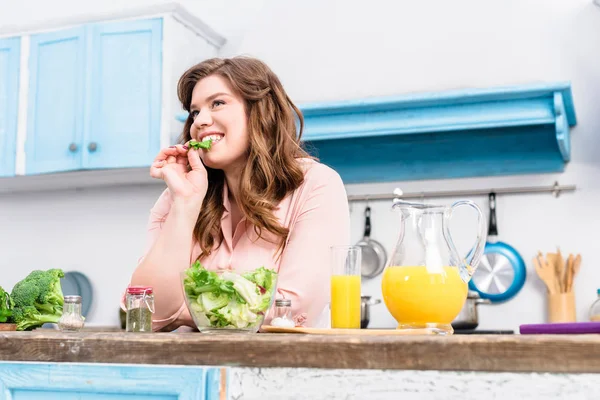 Übergewichtige lächelnde Frau am Tisch mit frischem Salat in der heimischen Küche — Stockfoto