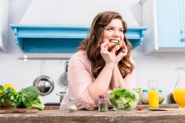 Übergewichtige lächelnde Frau am Tisch mit frischem Salat in der heimischen Küche — Stockfoto