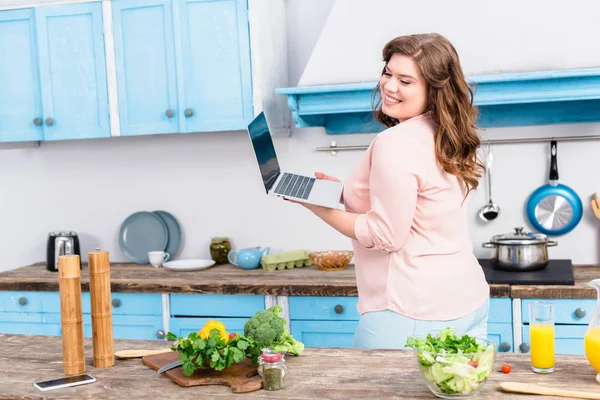 Femme souriante en surpoids avec ordinateur portable debout près de la table avec des légumes frais dans la cuisine à la maison — Photo de stock